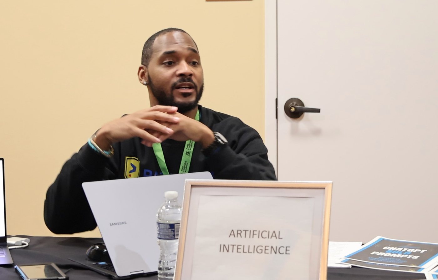 Jermaine Peguese at a table marked Artificial Intelligence, explaining with his hands